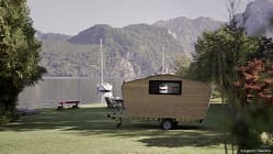 Der kleine Holz-Wohnwagen steht am Ufer eines Sees. Im Hintergrund segelt ein Boot vorbei und bewaldete Hügel säumen das Wasser.