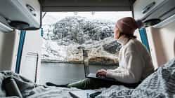 Shot of a mature woman working from the bed of her campervan. She pauses to take in the view of the loch and the snowy mountains