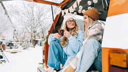 Two cheerful women friends sitting in a van in winter camp and having fun, enjoying holiday.