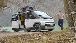 Kia PV5 Alpincamper mit ausgefahrenem Aufstelldach am Seeufer, ein Mann lehnt am Fahrzeug.