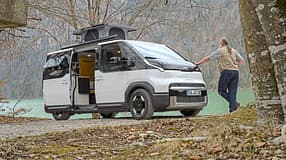 Kia PV5 Alpincamper mit ausgefahrenem Aufstelldach am Seeufer, ein Mann lehnt am Fahrzeug.