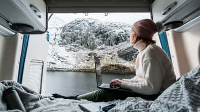Shot of a mature woman working from the bed of her campervan. She pauses to take in the view of the loch and the snowy mountains