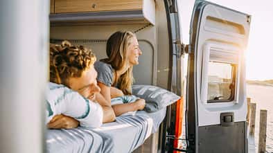 Zwei Personen liegen entspannt im Bett eines Sunlight Cliff 640 Campervans und blicken durch die geöffnete Hecktür auf eine sonnige Landschaft.