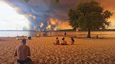 TOPSHOT - Beach goers rest on the beach of Moutchic on the shore of Lake Lacanau (Etang de Lacanau) in Lacanau , southwestern France on July 24, 2026, as clouds of smoke rise into the sky from the Gironde wildfire. The fire raging in the northern part of the Arcachon Basin in Gironde, southwest France, has swept through more than 3,400 hectares, according to the latest figures released by the prefecture. More than 20,000 residents and tourists have been evacuated from this part already devastated by flames in 2022. Scientists have long warned that climate change caused by human use of fossil fuels will make periods of drought more intense and longer-lasting, creating the ideal conditions for wildfires. (Photo by Maximilien LAMY / AFP via Getty Images)