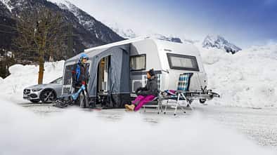 Das Foto zeigt eine Wintercamping-Szene auf dem Eurocamping Arlberg: ein Wohnwagen (Eriba) mit aufgebautem Vorzelt, davor zwei Personen in Winterbekleidung mit Ski- bzw. Schneesportgerät. Neben dem Caravan stehen Campingstühle und Ausrüstung, umgeben von hohen Schneewällen und einer alpinen Bergkulisse. Die Aufnahme verortet die Situation am Arlberg und betont Camping im Winterbetrieb.