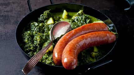 nobody,traditionally german,potato,gleaming,prepared,hearty,smoked,dish,ready to eat,minced pork sausage,north german,dark background,silver spoon,braising pan,pan Curly kale with potatoes and two minced pork sausages in pan