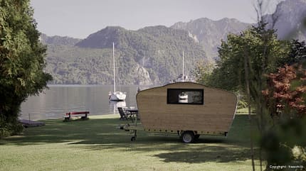 Mini-Wohnwagen, Teardrop, Nachhaltigkeit, Holz, Handarbeit, Natur Der kleine Holz-Wohnwagen steht am Ufer eines Sees. Im Hintergrund segelt ein Boot vorbei und bewaldete Hügel säumen das Wasser.