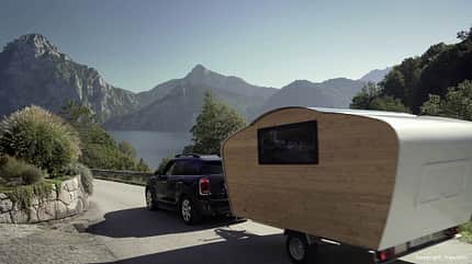 Mini-Wohnwagen, Teardrop, Nachhaltigkeit, Holz, Handarbeit, Natur Der Teardrop-Caravan Free Chilli hängt an einem Zugfahrzeug. Im Hintergrund ist ein See zu sehen, der von einem Gebirge eingerahmt wird.