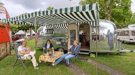 Airstream, Vordach, Campingtisch, Klappstühle, Thermoskanne, Blumen, Personen Airstream, Vordach, Campingtisch, Klappstühle, Thermoskanne, Blumen, Personen