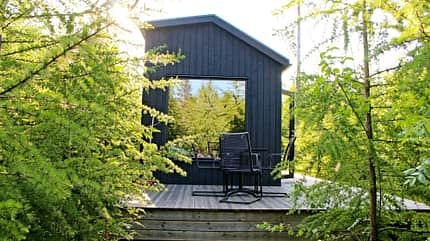 Schweden, Tiny House, Holzhaus, Waldhaus, Terrasse, Natur, Skandinavien, Ferienhaus, Minimalismus, Sommer Kleines, modernes Holzhaus in Schweden, umgeben von dichtem Grün und Bäumen, mit Terrasse und Stuhl.