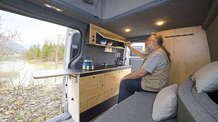 Innenaufnahme eines Kia PV5 Alpincamper mit ausgebautem Küchenmodul und einer Person, die ein Staufach öffnet.