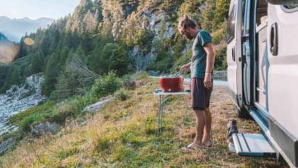 Man cooks on portable barbecue outside his van, mountain view