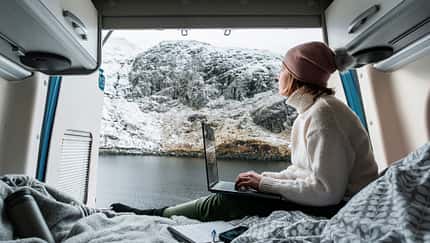 Shot of a mature woman working from the bed of her campervan. She pauses to take in the view of the loch and the snowy mountains