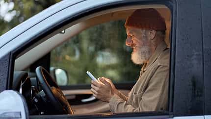 Happy older man traveler sitting in camper van using mobile in camping.