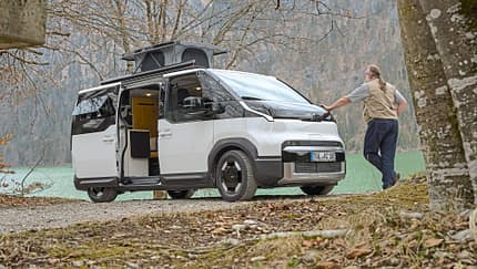 Kia PV5 Alpincamper mit ausgefahrenem Aufstelldach am Seeufer, ein Mann lehnt am Fahrzeug.