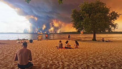 TOPSHOT - Beach goers rest on the beach of Moutchic on the shore of Lake Lacanau (Etang de Lacanau) in Lacanau , southwestern France on July 24, 2026, as clouds of smoke rise into the sky from the Gironde wildfire. The fire raging in the northern part of the Arcachon Basin in Gironde, southwest France, has swept through more than 3,400 hectares, according to the latest figures released by the prefecture. More than 20,000 residents and tourists have been evacuated from this part already devastated by flames in 2022. Scientists have long warned that climate change caused by human use of fossil fuels will make periods of drought more intense and longer-lasting, creating the ideal conditions for wildfires. (Photo by Maximilien LAMY / AFP via Getty Images)