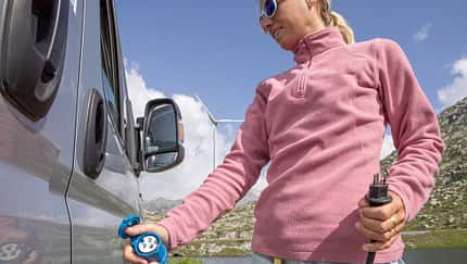 Camperin mit einem Adapter vom Schweizer Stecker auf den blauen CEE-Anschluss schließt ihr Wohnmobil an den Landstrom an. Woman plugging her camper van to recharge the battery, wind turbine in the background