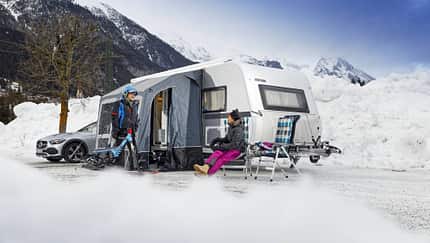 Das Foto zeigt eine Wintercamping-Szene auf dem Eurocamping Arlberg: ein Wohnwagen (Eriba) mit aufgebautem Vorzelt, davor zwei Personen in Winterbekleidung mit Ski- bzw. Schneesportgerät. Neben dem Caravan stehen Campingstühle und Ausrüstung, umgeben von hohen Schneewällen und einer alpinen Bergkulisse. Die Aufnahme verortet die Situation am Arlberg und betont Camping im Winterbetrieb.