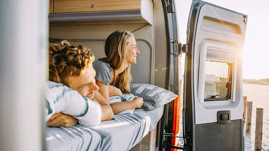 Zwei Personen liegen entspannt im Bett eines Sunlight Cliff 640 Campervans und blicken durch die geöffnete Hecktür auf eine sonnige Landschaft.