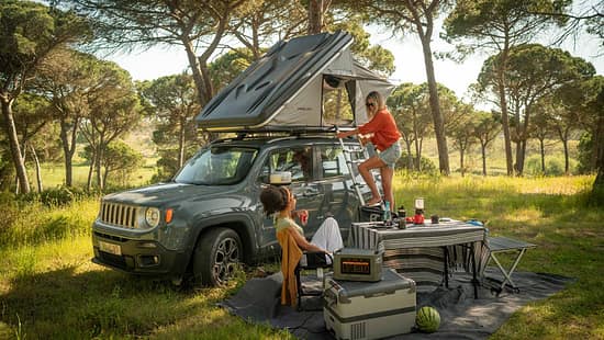 Zurück zur Natur: Prime Tech Delta Dachzelt zwischen hohen Bäumen, Leiter ausgeklappt, zwei Frauen genießen das Camping-Setup. Waldluft, Vogelgezwitscher und das Dach über dem Kopf – so schmeckt Freiheit.