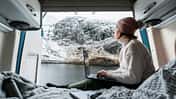 Shot of a mature woman working from the bed of her campervan. She pauses to take in the view of the loch and the snowy mountains
