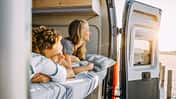 Zwei Personen liegen entspannt im Bett eines Sunlight Cliff 640 Campervans und blicken durch die geöffnete Hecktür auf eine sonnige Landschaft.
