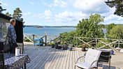 Terrasse mit Blick auf einen See in Schweden, ein Mann steht am Rand, Gartenmöbel und Blumentöpfe sichtbar.