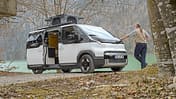 Kia PV5 Alpincamper mit ausgefahrenem Aufstelldach am Seeufer, ein Mann lehnt am Fahrzeug.